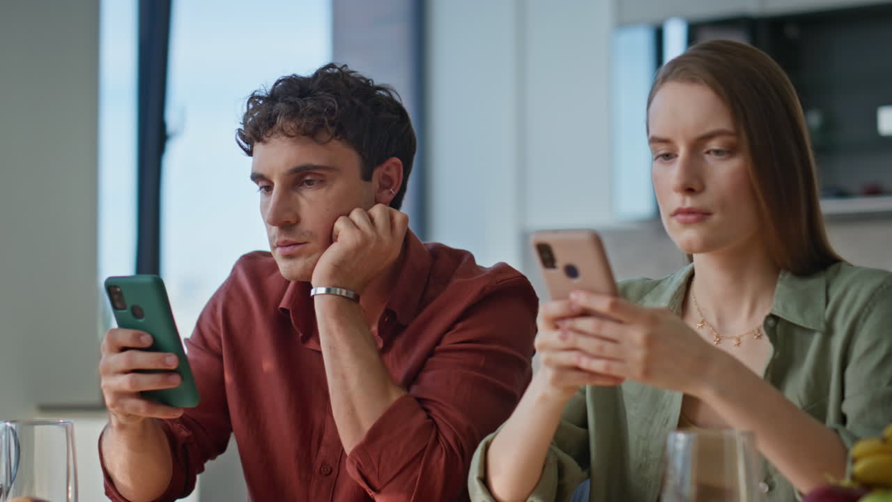 Serious couple involved cellphones sitting kitchen at home breakfast closeup