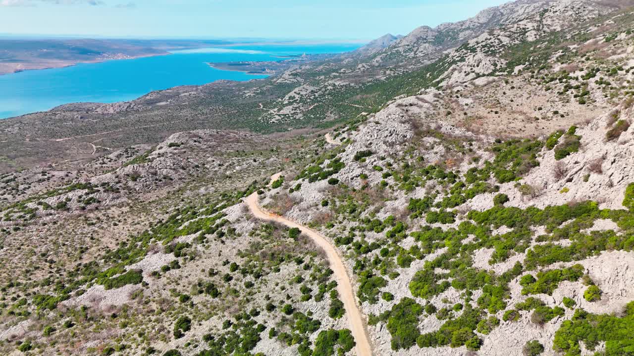 Vista drone DJI sul Monte Velebit, seguendo una strada sterrata scavata nei pendii fino al Mare Adriatico, ripresa sotto un sole splendente e in condizioni di cielo sereno.