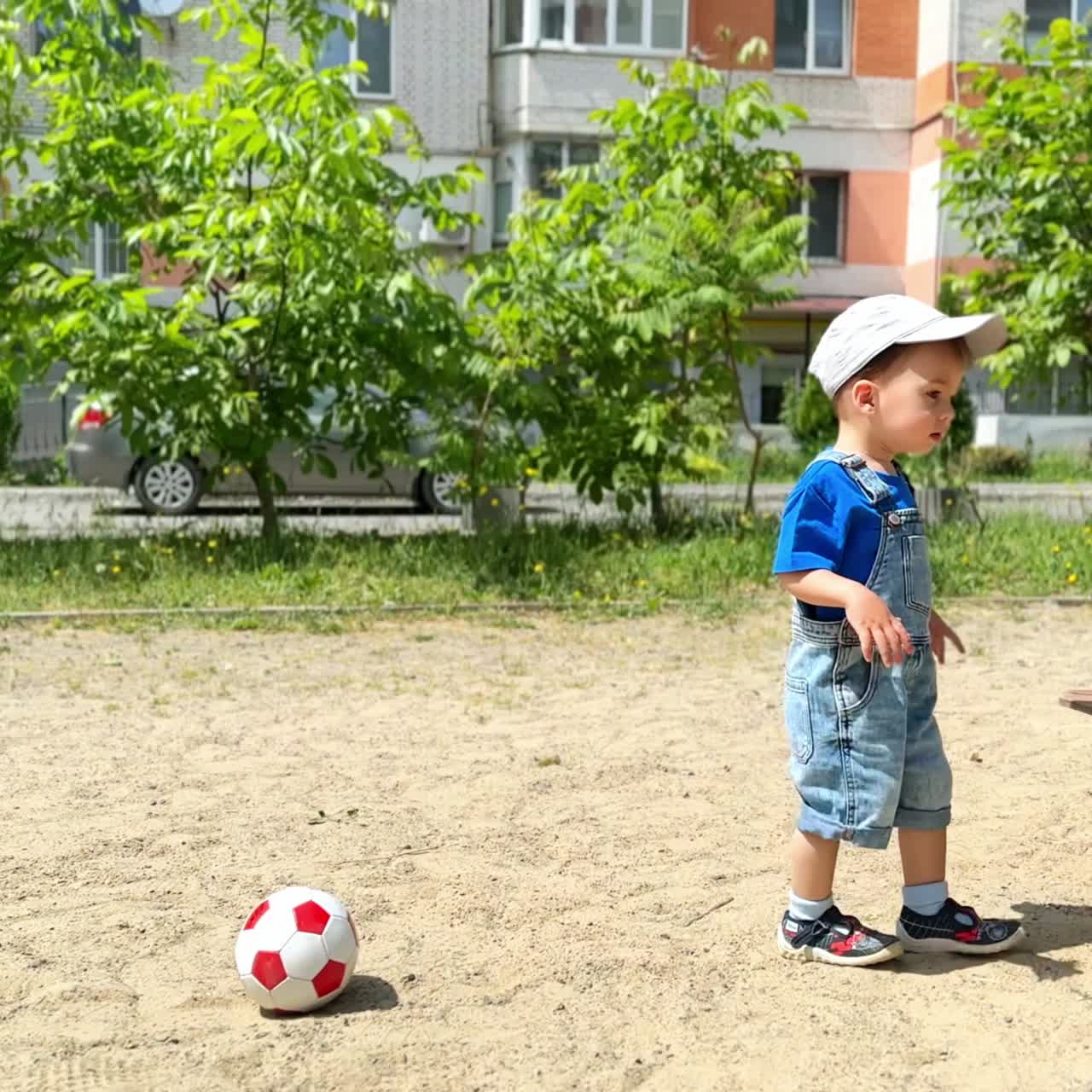 Caucasian baby boy in a cap comes up to a sandbox. Little toddler gets inside to play with sand