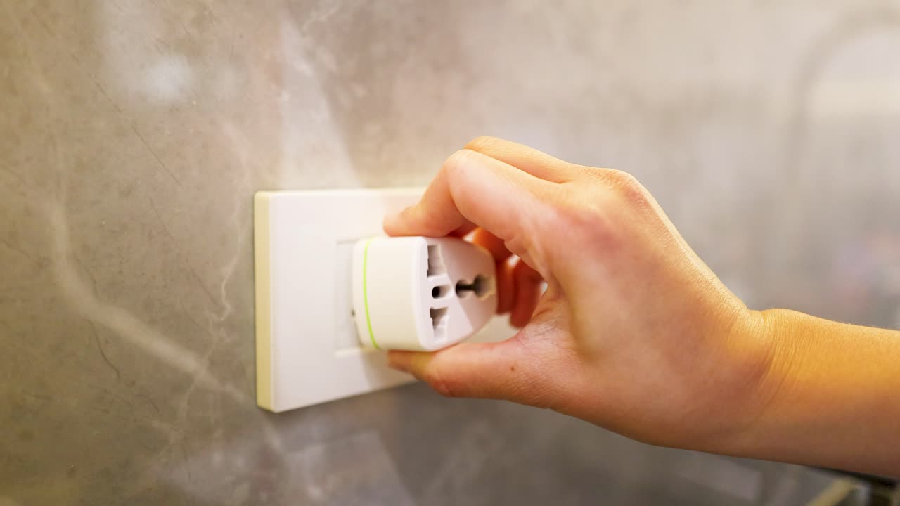 A close-up shot shows a hand plugging a white universal travel adapter into a socket