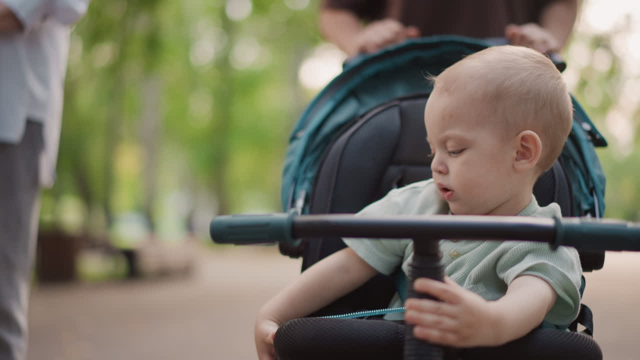 White toddler with pacifier exploring stroller bar and surroundings in park, curious gaze, caregiver hands visible, soft bokeh trees, intimate family lifestyle moment capturing small gestures