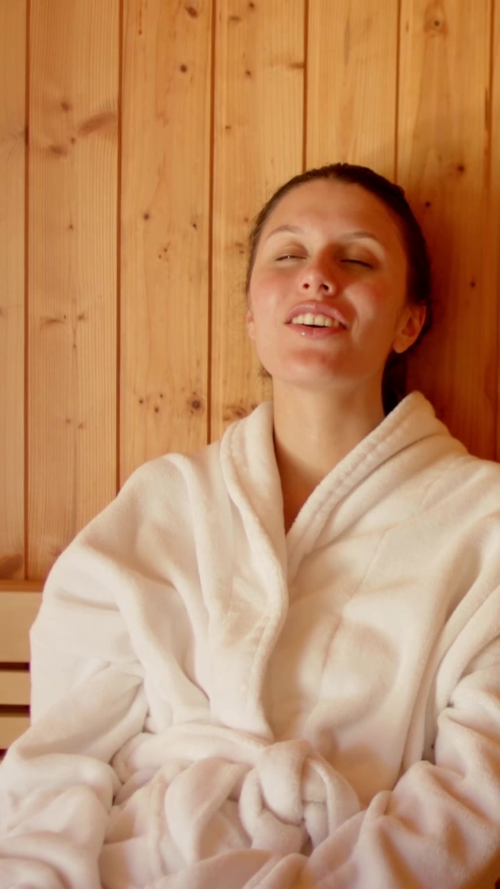 Vertical video: Sitting woman in robe relaxing opening eyes smiling on bench by wood wall robe knot