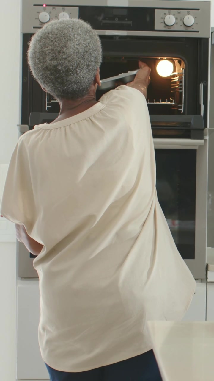 Video verticale: Anziana con top color crema che, illuminata dalla luce del forno, posiziona una teglia per muffin usando un canovaccio da cucina, mentre inforna.