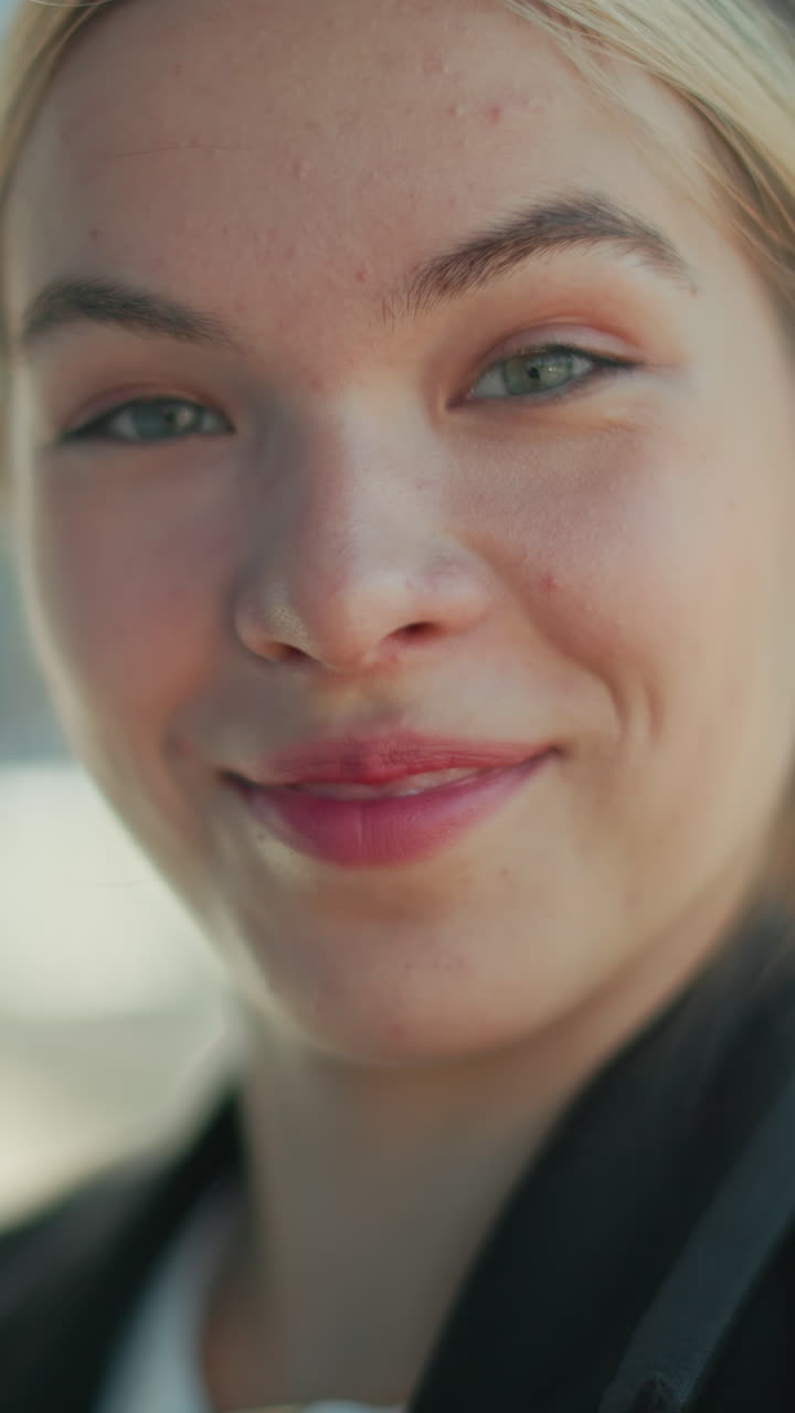 Lady in professional setting operating phone stares at camera with surprised look and subtle smile, dressed in formal outfit, standing outdoors in urban environment with soft blur city background