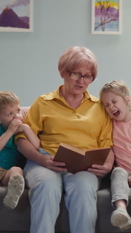 cute old lady with glasses reads book to children at night, the children listen to fairy tales with interest, sit on gray sofa with blanket in cozy bright room, the child yawns and wants to sleep