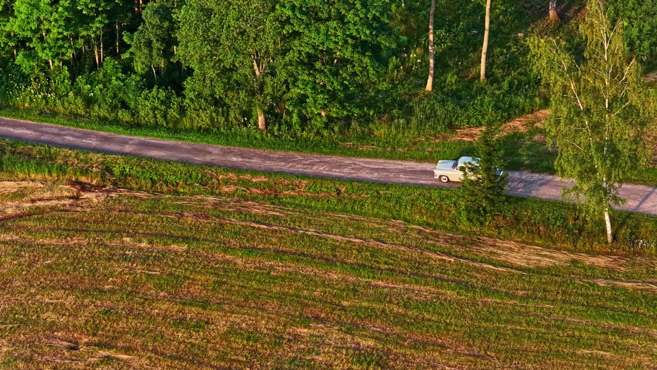 An aerial drone tracks an old, classic Soviet-era car as it drives along a scenic country road through forests and fields in Latvia during a beautiful golden hour sunset