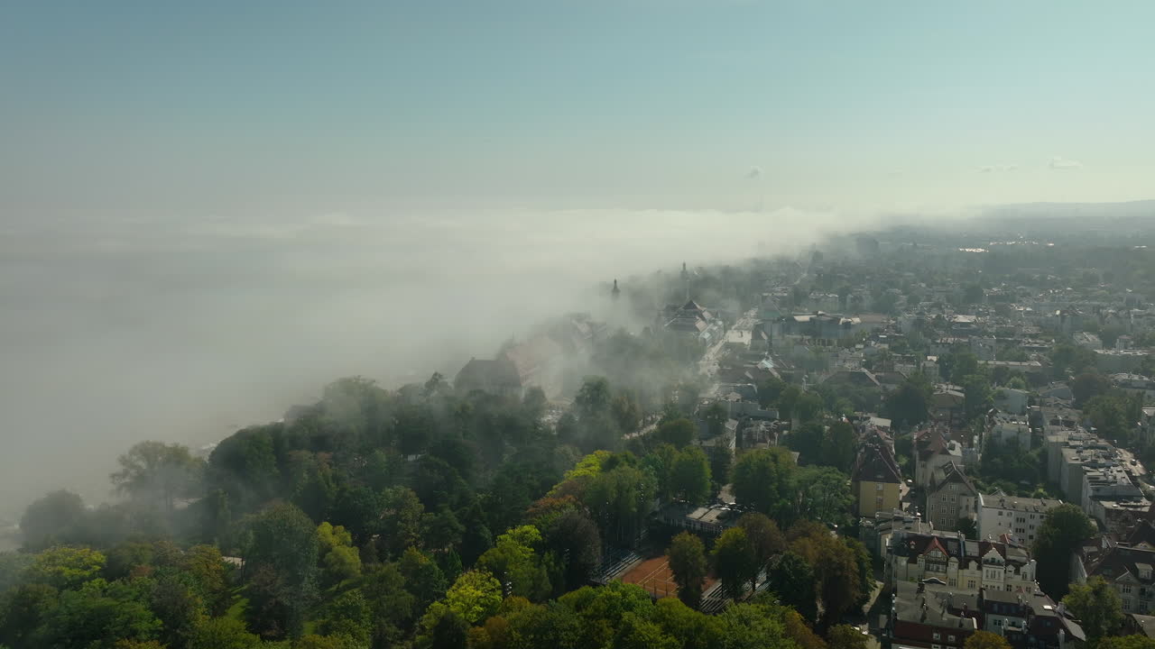 Aerial shot of Sopot shrouded in fog, with lush trees and urban buildings emerging through the misty landscape.