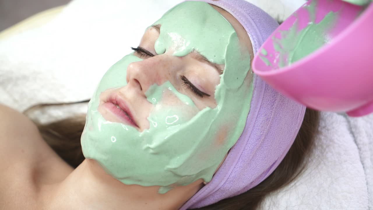 Facial mask on face. Therapist applying face mask to a beautiful young woman in spa using cosmetics brush