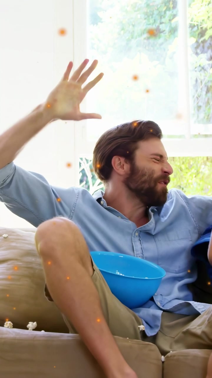 Vertical video: Dad lifting popcorn bowl as entertainment, son reacting, kernels spilling on couch