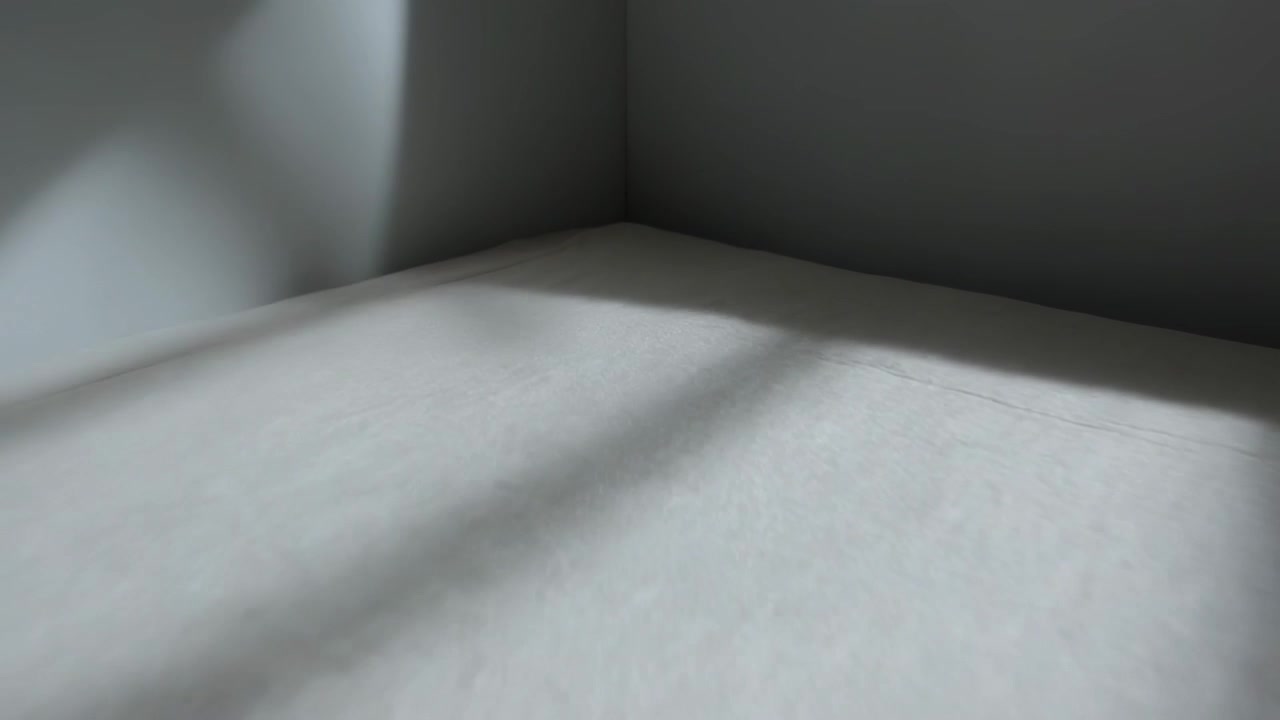 Telecamera in movimento che rivela un materasso con lenzuola in una camera da letto, esponendo ombre di finestre e pieghe.