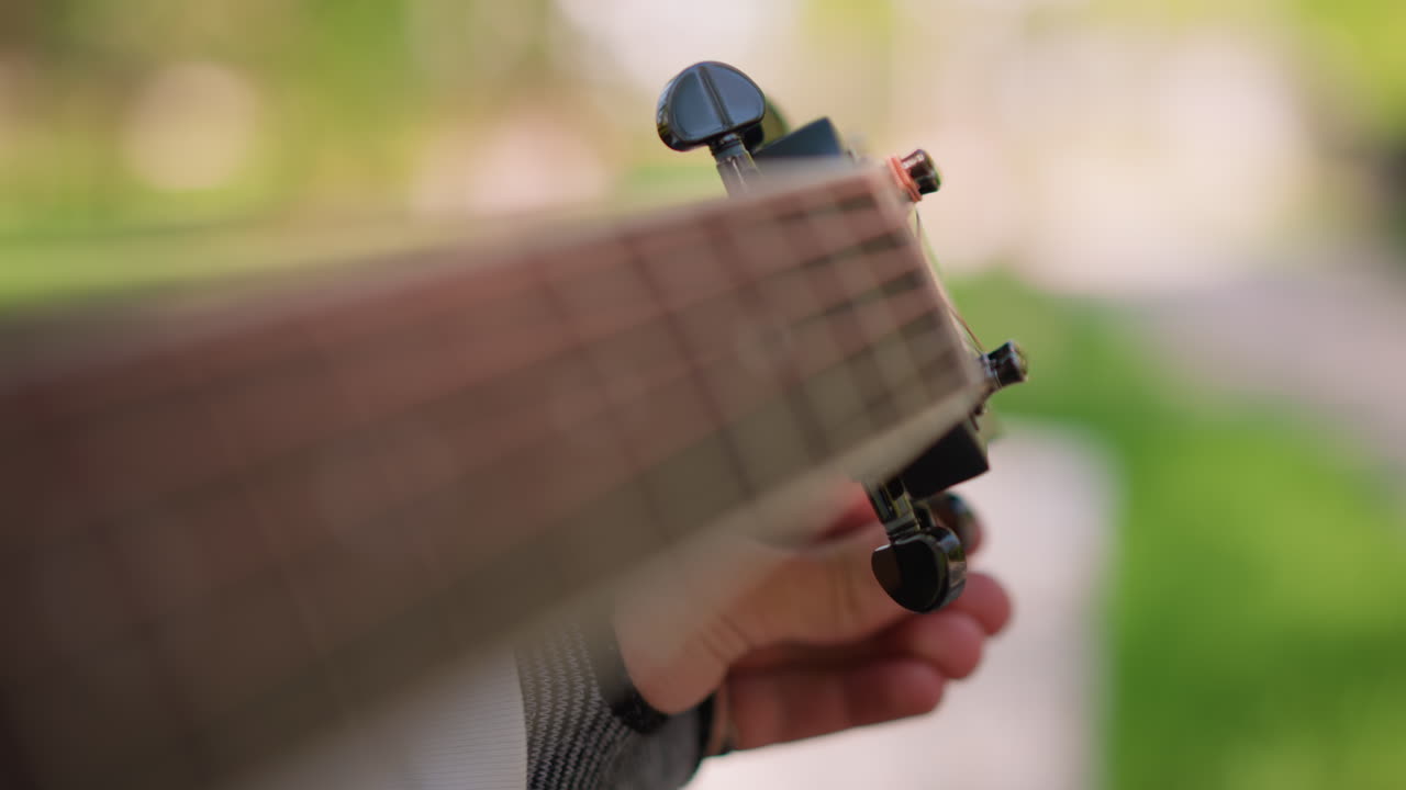 outdoor closeup hands tuning acoustic guitar headstock wrist brace fingerless glove soft sunlight bokeh grass park musician practicing gentle adjustment preparing performance focused technique