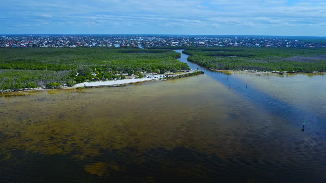 Le zone umide costiere e un corso d'acqua tortuoso separano una spiaggia sabbiosa e un'area di parcheggio da una vasta area urbana in Florida, sotto un cielo nuvoloso che copre il paesaggio