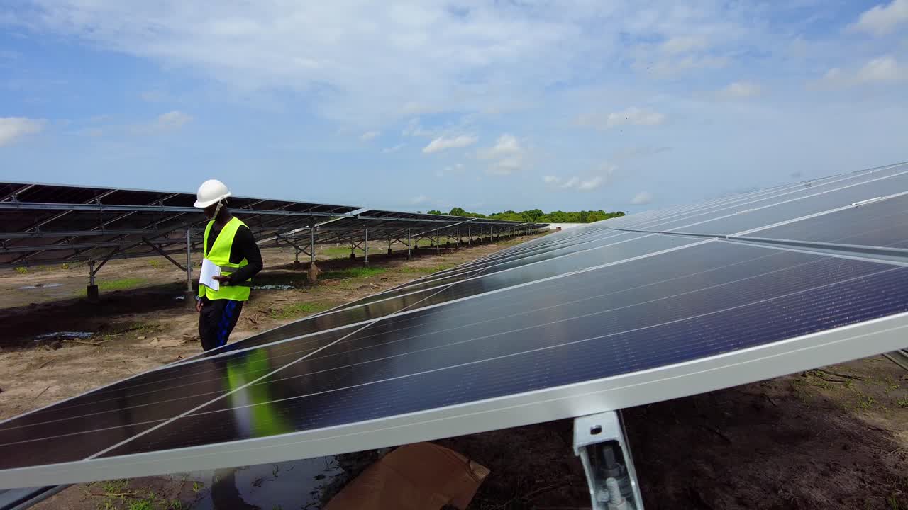 ingeniero africano negro caminando en una granja de energía renovable de paneles solares con casco protector llevando un cuaderno de papel con datos de mediciones de la eficiencia de la planta fotovoltaica en áfrica