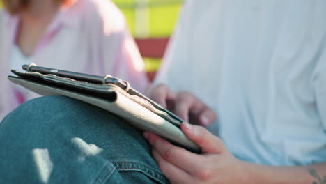 tech-savvy male types on his tablet, leg crossed at the knee, seated outdoors with a blurred view of someone in a pink top close by, greenery in the background