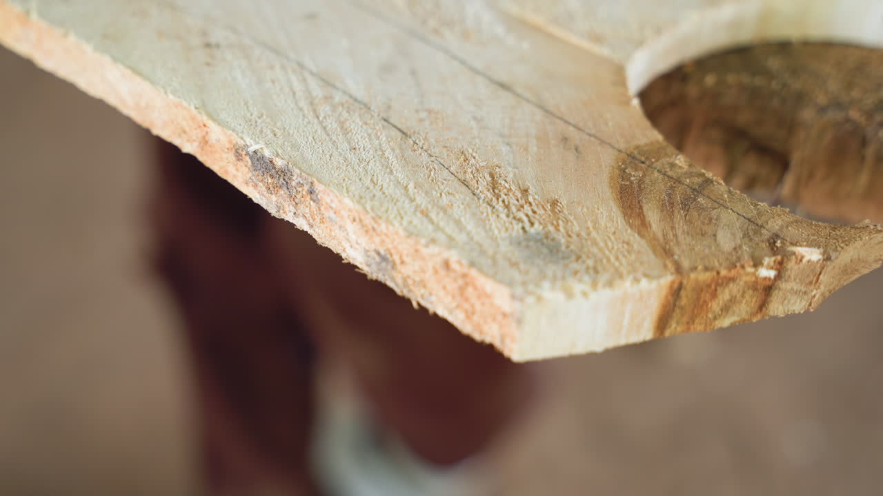 Vista angolata di un bordo di legno curvo con intaglio circolare, fibre grezze e segatura lungo il perimetro, falegname sfocato sullo sfondo, calda atmosfera dell'officina, tavola ispezionata per l'adattamento e la finitura finale.
