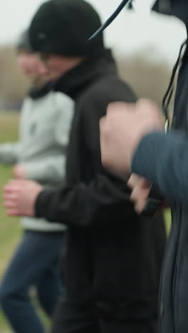 close-up of three people jogging on a track near a grassy field, with a clear view of the adult wearing a whistle in the foreground and a blurred view of the other young boys in the background