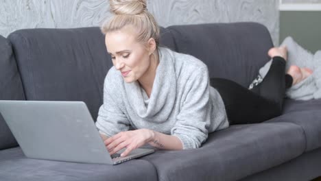 glückliche blonde frau, die auf dem sofa liegt und am laptop arbeitet