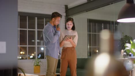 Focused-diverse-male-and-female-colleague-standing-talking-and-using-tablet-at-night-in-office