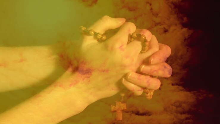 Animation of hands of person praying and holding a Christian rosary with cross over orange clouds