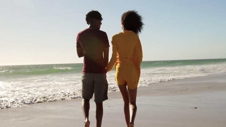 Di spalle, coppia afroamericana che tiene in mano una bottiglia di birra e cammina insieme sulla spiaggia 4k