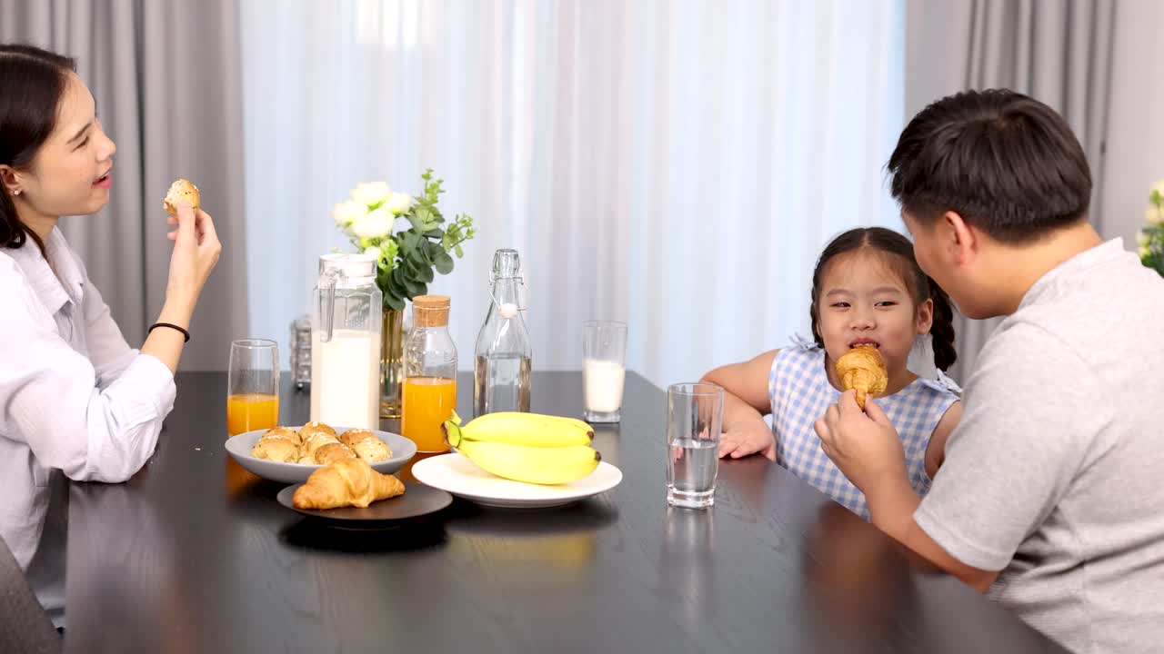 Asian family enjoys breakfast with croissants and juice at modern dining table in daylight