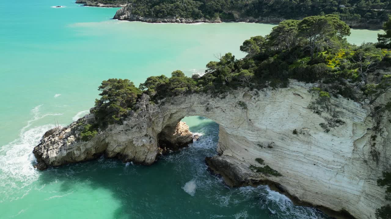 Stunning 4K drone footage of a natural sea arch, rugged limestone cliffs, turquoise waters, lush coastal vegetation along the Gargano Peninsula in Puglia, southern Italy