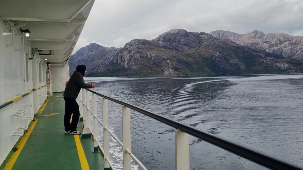 Uomo che si gode un viaggio in traghetto attraverso i fiordi selvaggi della Patagonia cilena, camminando accanto alle ringhiere con imponenti montagne e acque a specchio sullo sfondo.