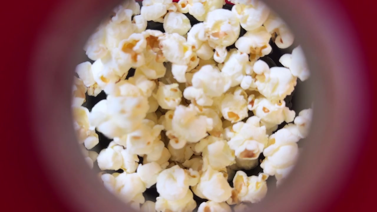 Ripresa macro in soggettiva attraverso un'apertura circolare di popcorn appena scoppiati all'interno di una macchina rossa. L'illuminazione brillante enfatizza la consistenza croccante e bianca e i chicchi salati per un aspetto da snack cinematografico.