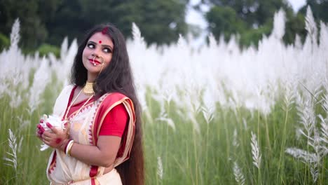 a beautiful and religious married indian woman holding a shankha or conch shell looks behind in a field of white flowered grass or kaash phool