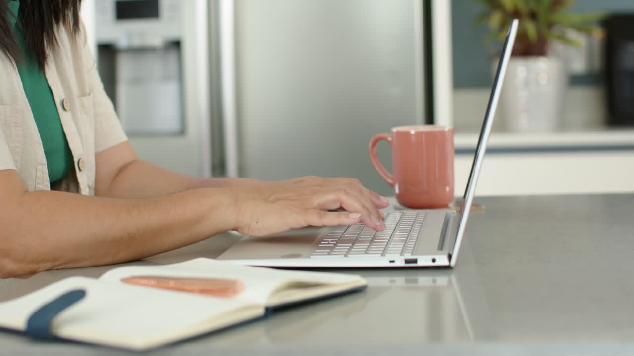 After pausing slightly, adult woman is typing on silver laptop at kitchen counter, using notebook