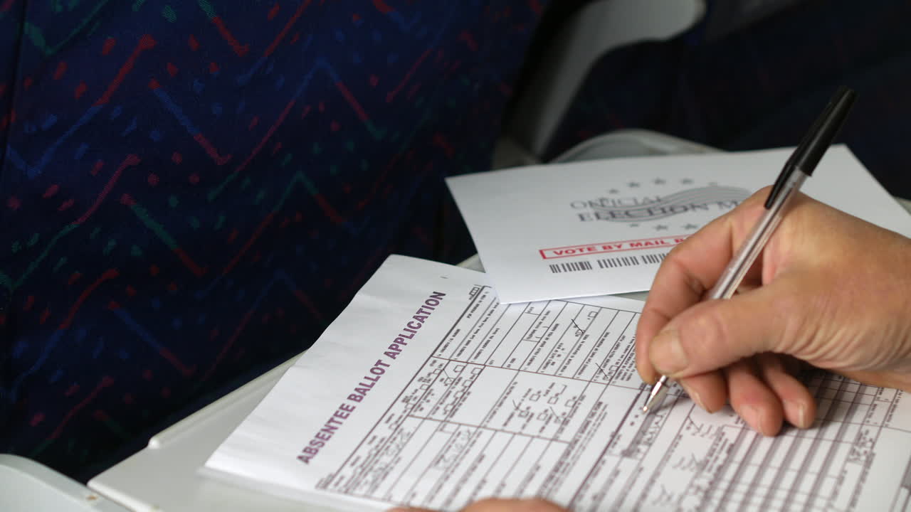Filling out an absentee ballot application with a pen while seated on an airplane. The "Official Election Mail" envelope is nearby, highlighting the process of voting by mail while traveling