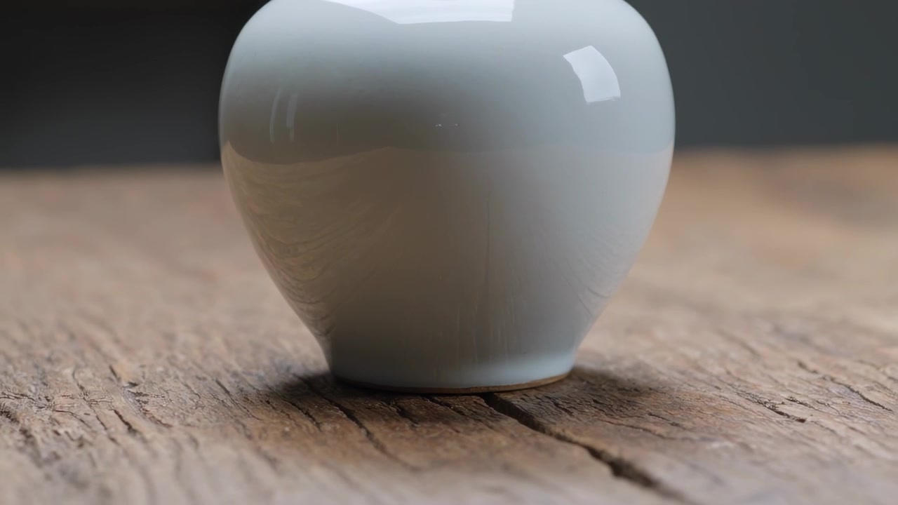A câmera se afasta revelando um vaso de cerâmica branco sobre uma tábua de madeira envelhecida, alinhando uma rachadura central.