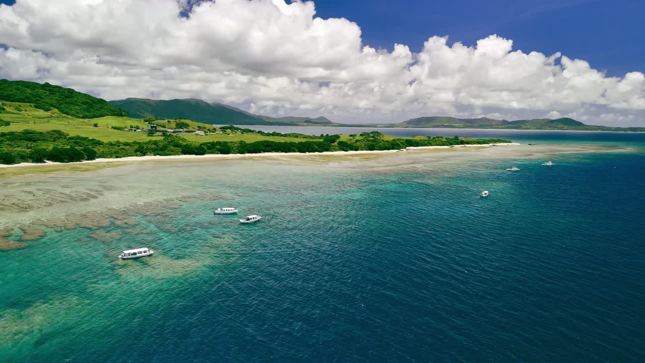 A gentle, cinematic aerial pan reveals a stunning tropical island paradise. Boats rest in the clear turquoise water above a coral reef, with lush green hills under a beautiful blue sky