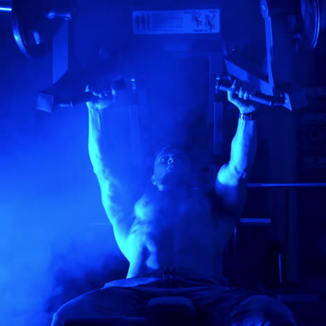 sportsman training on simulator. shirtless athlete doing his workout in the sports club. handsome bodybuilder in the gym. blue light filter and smoke.