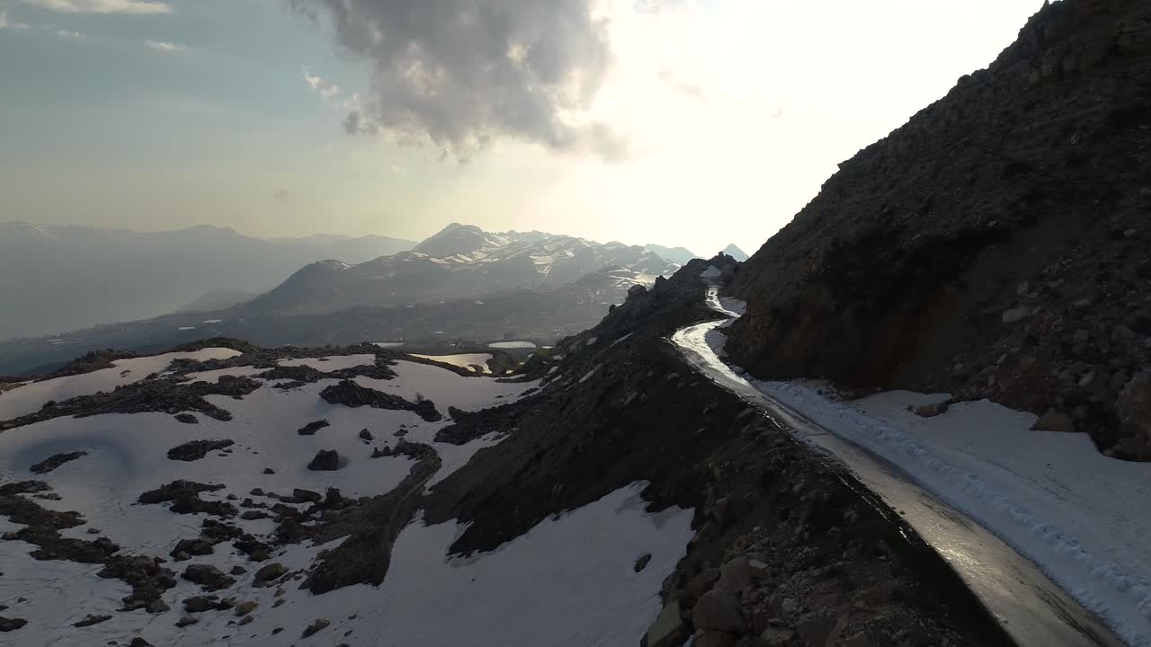 Aerial drone flight toward icy road on edge of rocky mountainside toward mountain range and sun