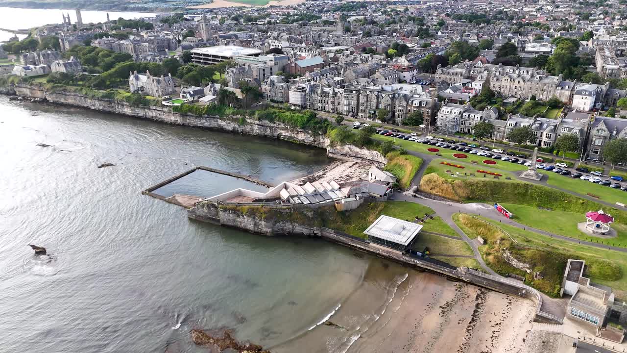 Drone footage captures the St Andrews coastline, tidal pools, and adjacent town under bright daylight, with smooth camera movement and wide scenic perspective