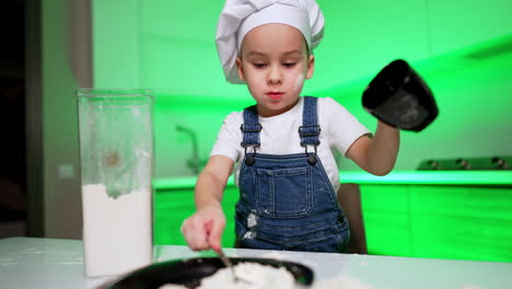 small baby boy in chef cap pours flour from the cup into the plate. caucasian toddler cooking in the kitchen.
