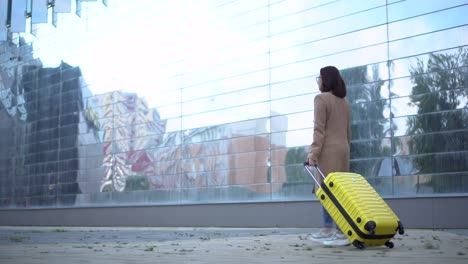 a young woman walks with a yellow suitcase. a girl with glasses and a coat walks along the mirrored building.