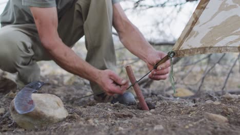 caucasian male survivalist attaching tent to stick peg at camp in wilderness