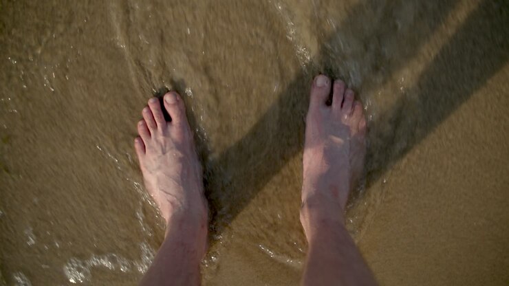 Gambe di giovane uomo su una spiaggia sabbiosa, con un'onda che si infrange.