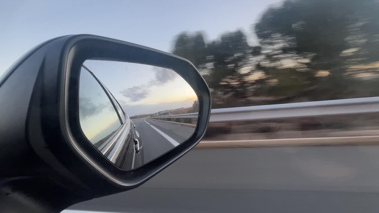 A car's rear view mirror shows a road with a sunset in the background