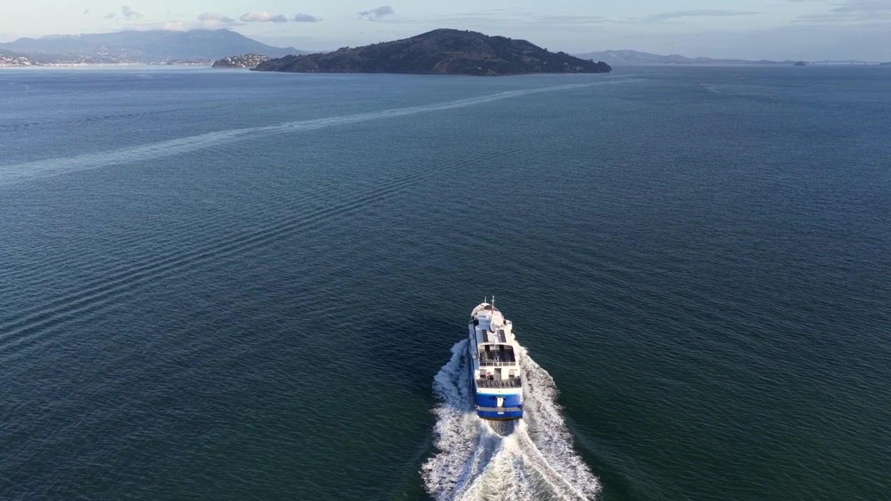Barco de pasajeros navegando por la Bahía de San Francisco, dejando una estela blanca, con aguas abiertas y una isla a lo lejos bajo una suave luz de verano. Escena de viaje marítimo pintoresca en Estados Unidos. Seguimiento aéreo.