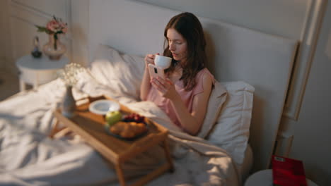 calm girl enjoying breakfast drinking coffee in bed. relaxed female surfing web