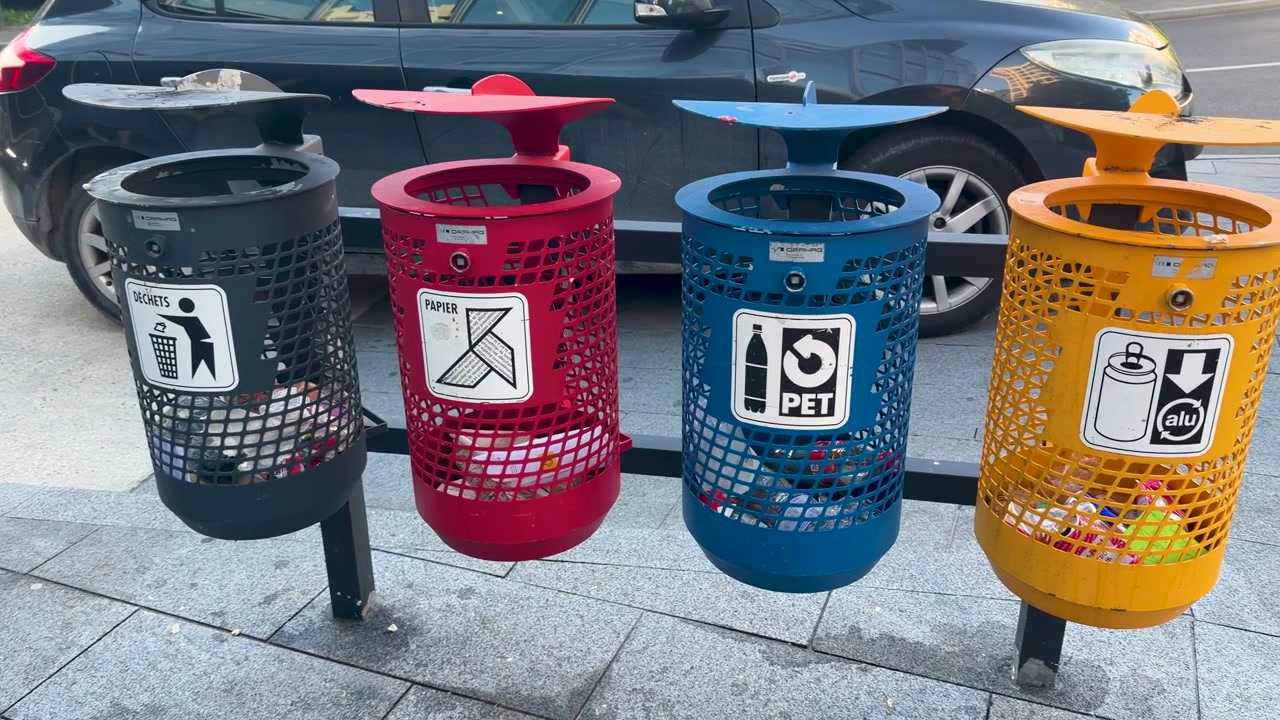 A slow panning shot reveals four color-coded recycling bins for sorting waste on a sidewalk.