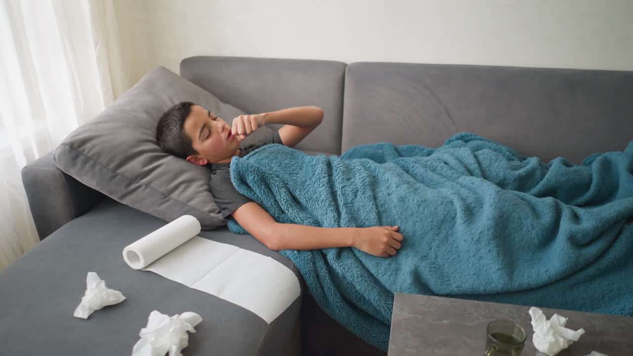 Aerial view of scattered room with child lying on couch adjusting under blanket, drug thermometer and medicines placed on table, tissues scattered around