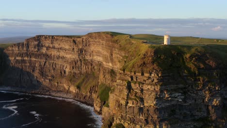 Aerial-rising-shot-reveals-the-Cliffs-of-Moher-wall-lit-by-sunset,-and-Irish-fields,-County-Clare