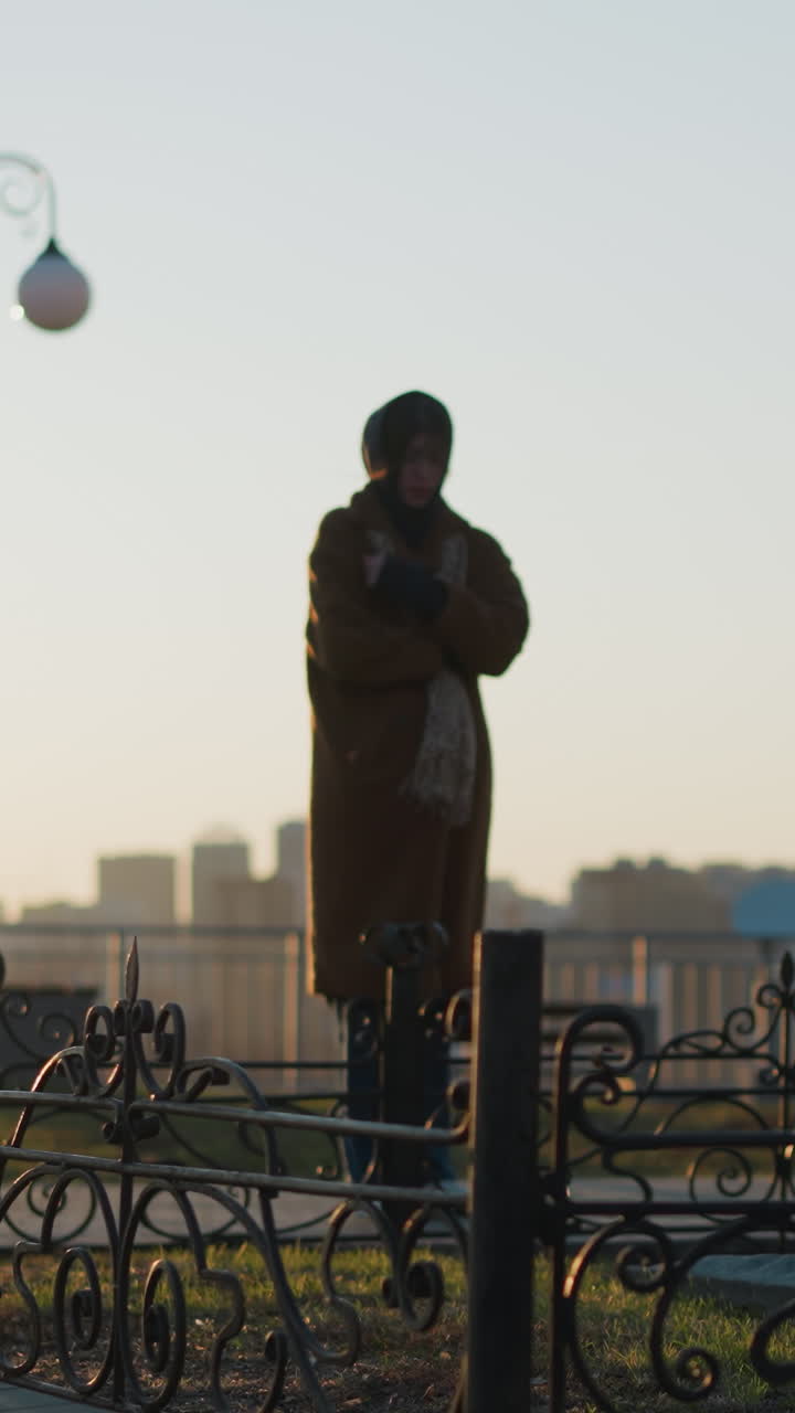 A woman wearing a coat and a scarf stands in front of a stone monument in a park during sunset. The scene is contemplative, with the warm light of the setting sun casting long shadows