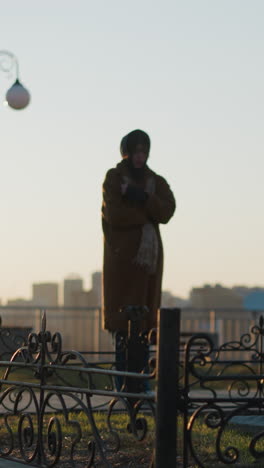 a woman wearing a coat and a scarf stands in front of a stone monument in a park during sunset. the scene is contemplative, with the warm light of the setting sun casting long shadows