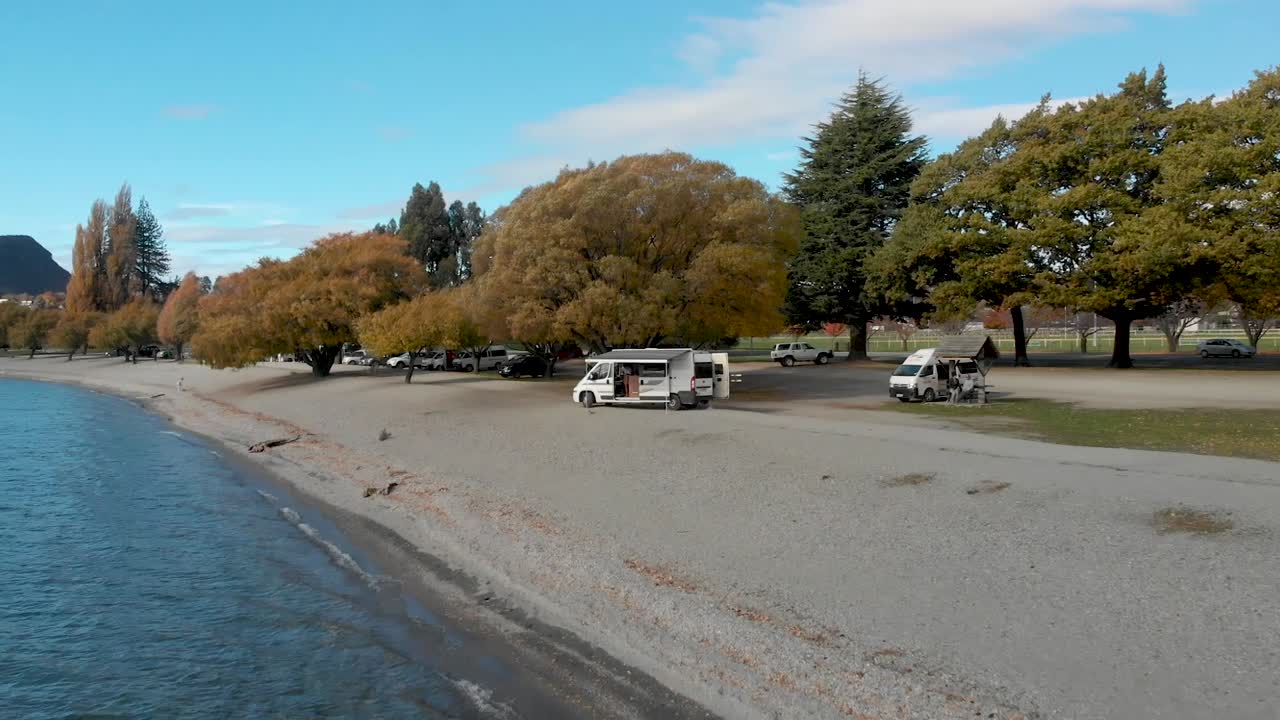 Aerial Drone flying towards a campervan on a beach above beautiful blue lake in Lake Wanaka, New Zealand in autumn