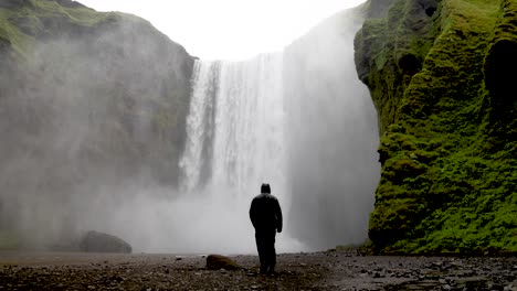 Skogafoss-Wasserfälle-In-Island-Mit-Mann-In-Regenjacke,-Der-Auf-Die-Wasserfälle-Zugeht